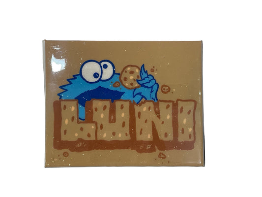 Decorative sign with a blue creature holding a cookie and the word 'Luna' on a beige background.